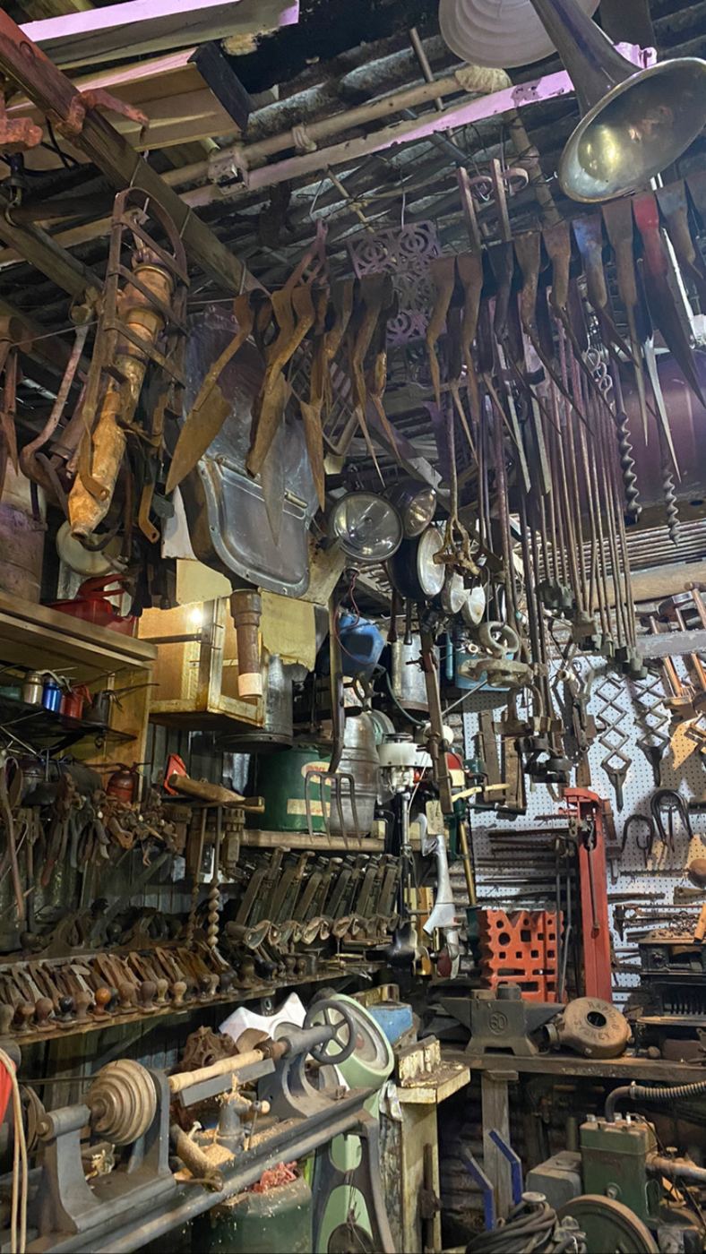 Hay knives, augers, car headlamps and lanterns hanging from the shed rafters above shelves of hand planes and vices